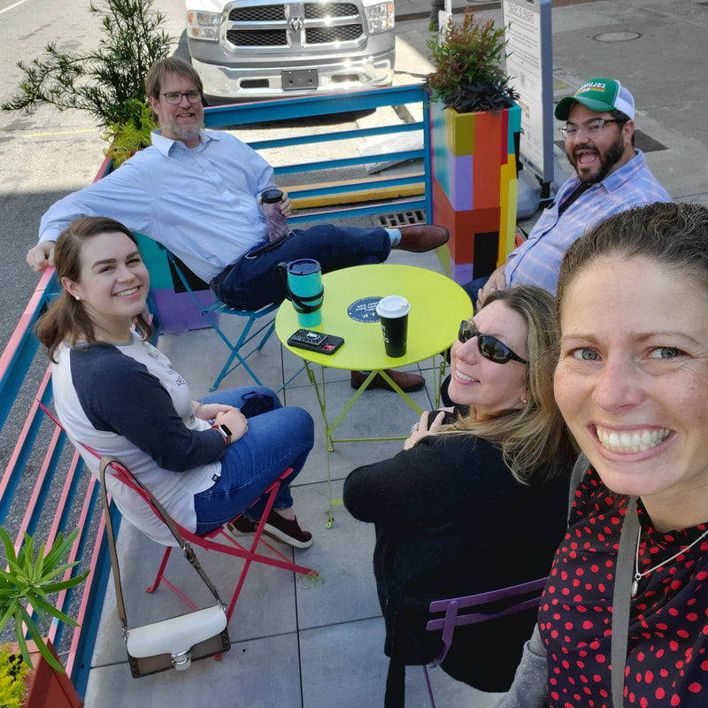 Photograph of a woman taking a group selfie in the Parklet on Washington.  She stands, smiling, in the lower righthand corner, as two women and two men sit in brightly colored chairs and at a bright green table, also located within the parklet, and smiling at the camera.