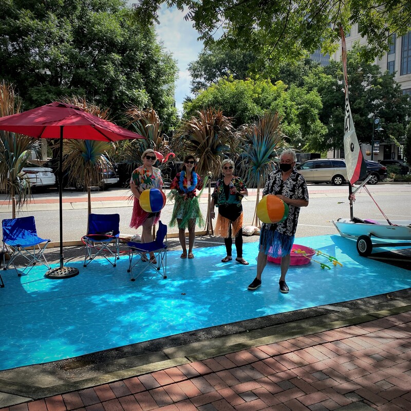 Photograph of people standing in a parklet on Main Street, on top of a blue plastic made to look like water.  A patio umbrella, palm fronds, and a sailboat surround them.  The people are wearing Hawaiian shirts and hula skirts and holding beach balls.